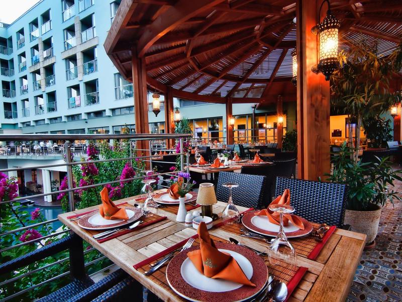 Cozy outdoor restaurant seating with wooden tables, orange napkins, and surrounding flowers near a hotel.
