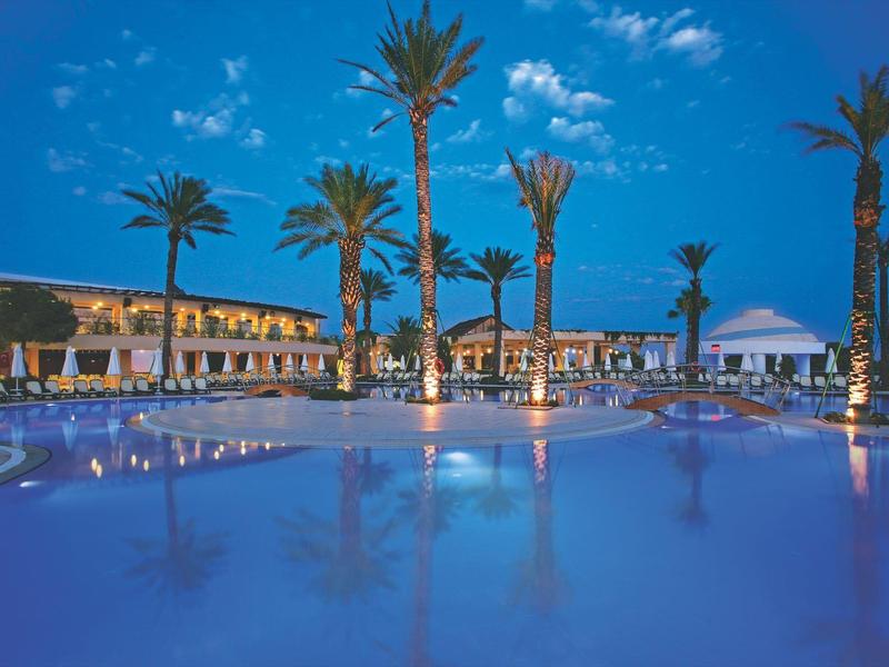 Luxury hotel pool area at dusk with palm trees and lit buildings reflecting on water.