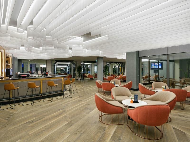 Spacious hotel lounge with modern bar, curved red chairs, wooden flooring, and stylish ceiling design.