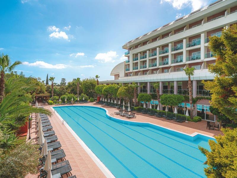 Grand hôtel avec piscine, chaises longues et parasols sous un ciel bleu.