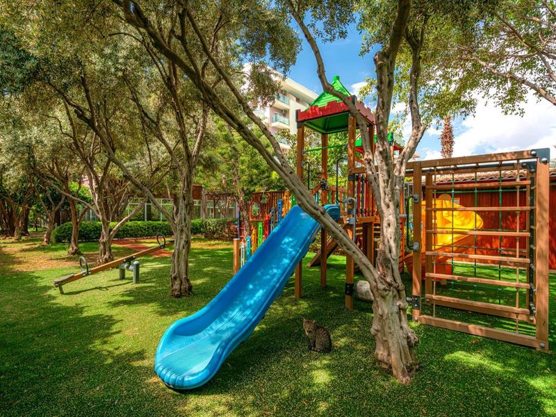 Aire de jeux pour enfants avec toboggan bleu et structure d'escalade dans un jardin verdoyant près d'un hôtel.