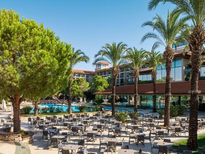 Venkovní prostor hotelu s mnoha stoly a židlemi pod palmami a bazénem v pozadí