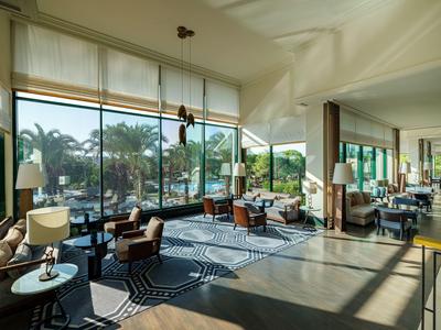 Světlý moderní hotelový salonek s velkými okny a výhledem na palmy.