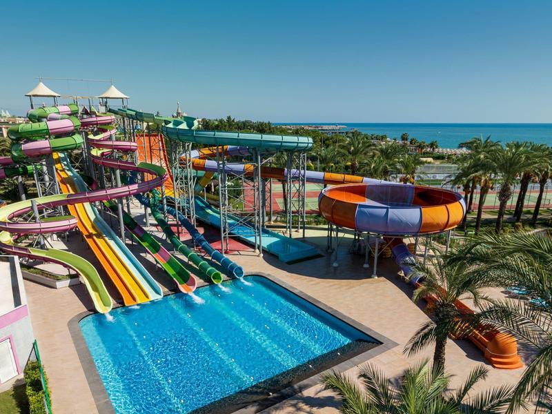 Barevný vodní park s několika tobogány a bazénem, obklopený palmami a mořem v pozadí.