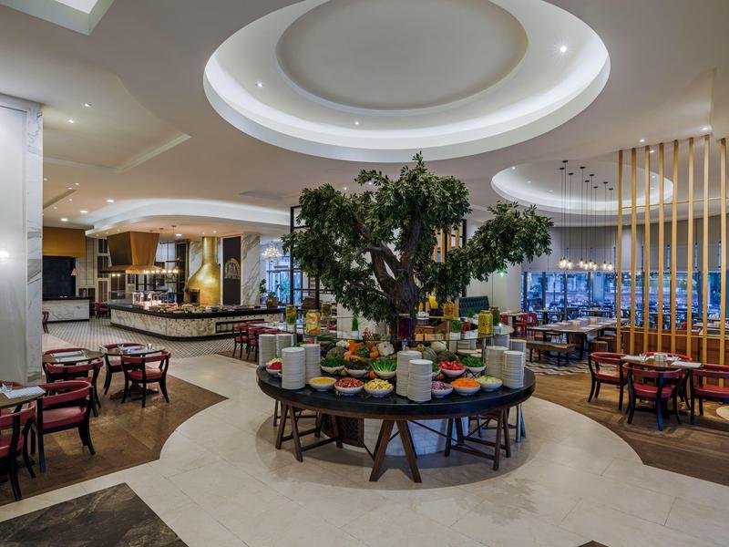 Moderní prostor hotelové restaurace s centrálním stromem a stolem s nabídkou snídaní.