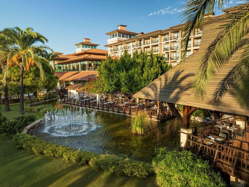 Hotelový komplex se rybníčkem, fontánou, palmami a venkovní restaurací pod modrou oblohou.