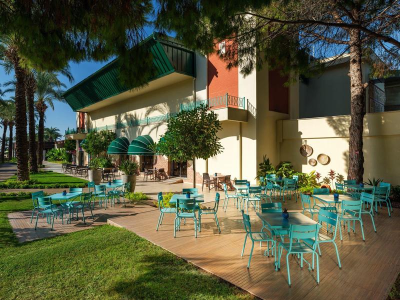 Venkovní prostor hotelu s modrými židlemi, stoly a zelenými slunečníky na terase vedle stromů.