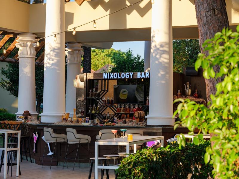 Unikátní mixology bar s bílými sloupy a venkovním posezením pod zeleným listím.