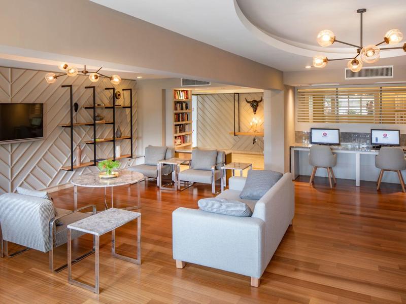 Area soggiorno moderna con divani, poltrone, TV e postazioni di lavoro in tonalità calde di legno.