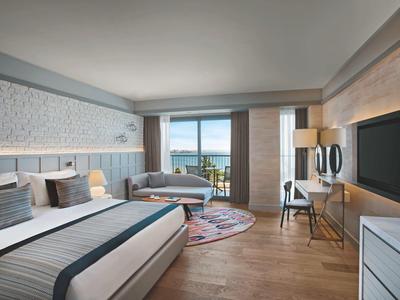 Camera d'hotel luminosa con due letti, scrivania, TV e balcone con vista mare.