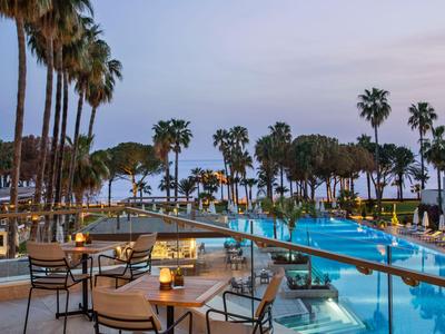 Terrazza serale con tavoli e sedie accanto a una piscina illuminata e palme sul mare.