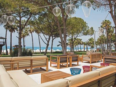 Lounge all'aperto con divani chiari e tavoli in legno sotto gli alberi con vista sul mare.