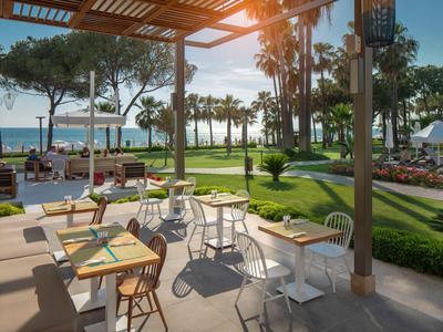 Terrazza esterna soleggiata di un hotel con tavoli, sedie e vista sul giardino e sul mare.