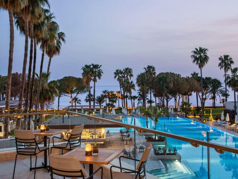 Area esterna moderna con piscina, posti a sedere e palme vicino al mare al tramonto.