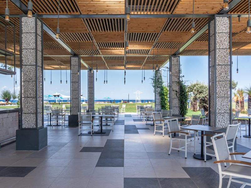 Open hotel pavilion with white chairs, wooden roof, and view of water and palm trees.