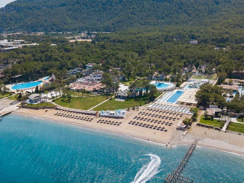 Hotel complex with pools, sun loungers, and clear sea in front of a forested mountain.