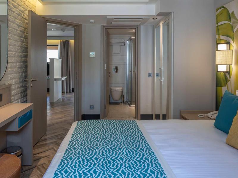 Modern hotel room with double bed, desk, and bathroom in the background.