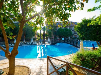 Ein sonniger Hotelpoolbereich mit blauem Wasser, Liegestühlen und Bäumen im Vordergrund.