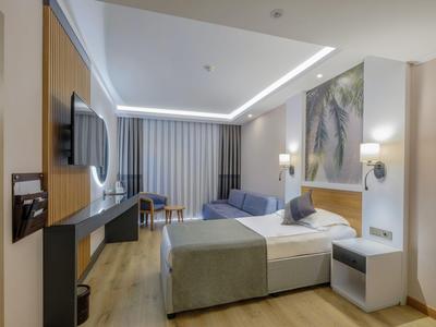 Modern eingerichtetes Hotelzimmer mit Doppelbett, Sofa, TV und Schreibtisch, helle Farben.