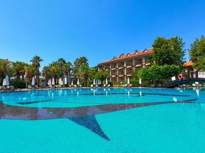 Großer Hotelpool mit klarem Wasser, umgeben von Sonnenschirmen und grünen Bäumen unter blauem Himmel.