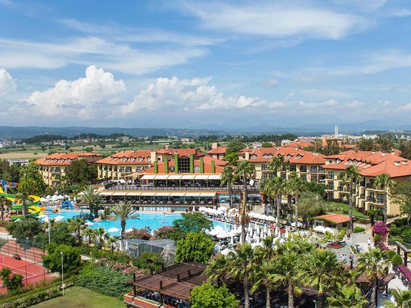 Large resort with multiple pools, red-roofed buildings, palm trees, and tennis courts under a blue sky.
