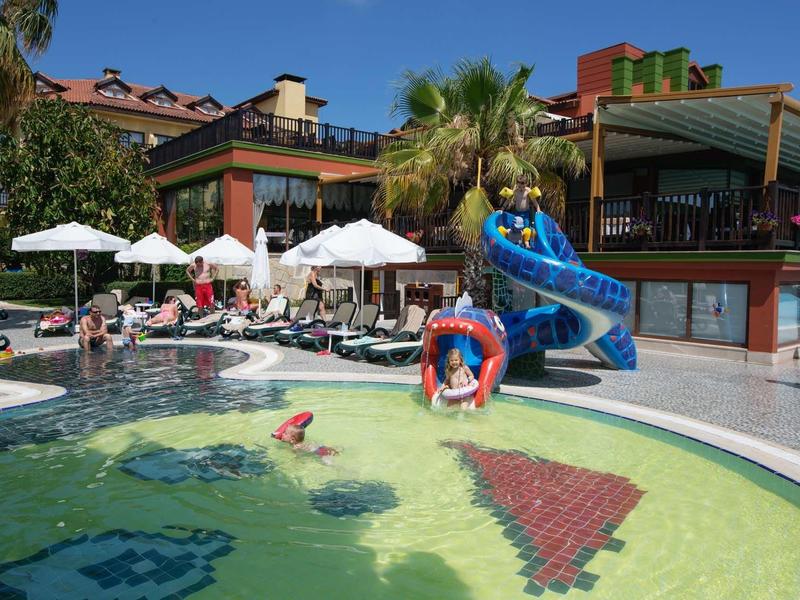 Outdoor pool with children playing, lounge chairs, umbrellas, and a water slide at a hotel.