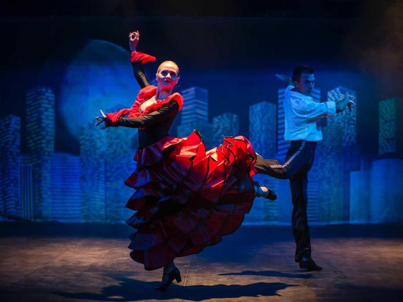 Dancers perform flamenco on stage with a vibrant red dress and dark background.