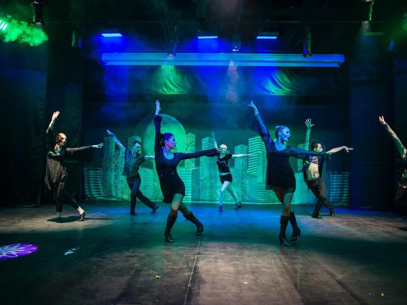 Dancers perform on stage under blue and green lighting with dynamic poses.