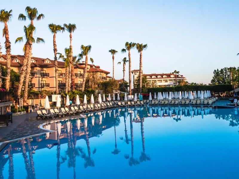 Resort pool with palm trees and sun loungers under clear blue sky.