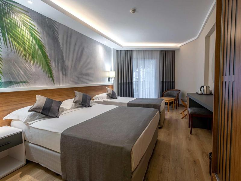 Modern hotel room with two beds, bedside tables, wooden flooring, and a large window with curtains.