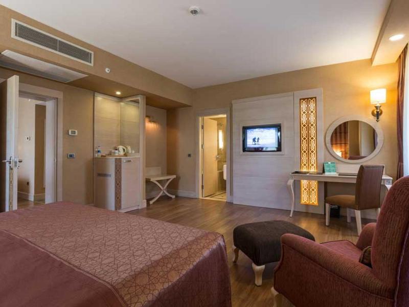 Modernes Hotelzimmer mit braunem Bett, Sessel, Schreibtisch, Spiegel und TV an der Wand.
