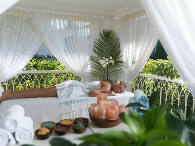 Ein heller Wellnessbereich mit weißem Vorhang, Liegen und Spa-Utensilien auf einer Veranda.