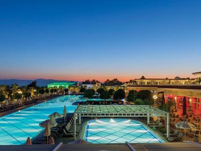Großer beleuchteter Poolbereich mit Hotelgebäuden und Palmen bei Sonnenuntergang.