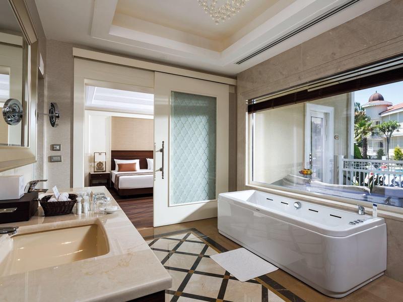 Modern bathroom with a large bathtub and double sink, connected to a bedroom through a glass door.