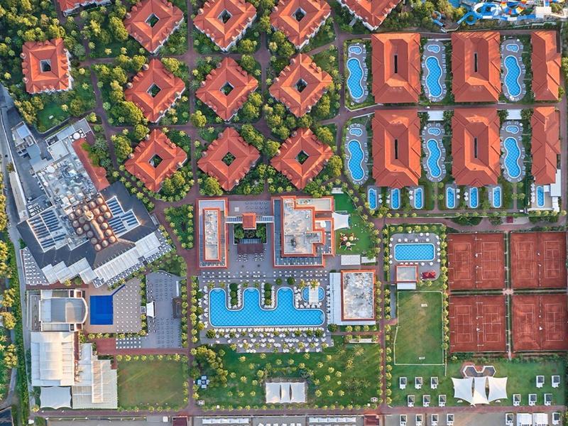 Aerial view of a resort with red-roofed buildings, pools, tennis courts, and beach access.
