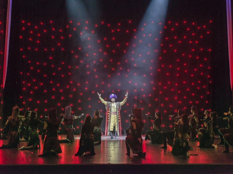 Stage performance with a central figure illuminated by spotlight, surrounded by dancers in dark costumes.