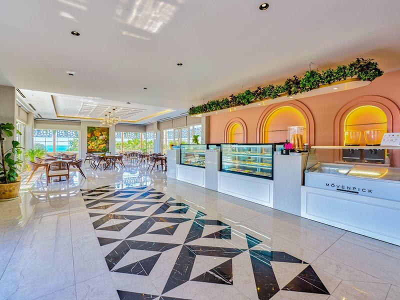 Bright hotel lobby with patterned floor, seating area, and a colorful reception desk with plants.