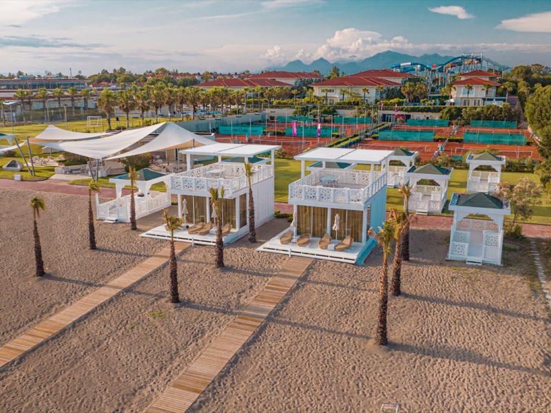 Modern beachfront villas with white roofs on sandy beach and mountainous background.