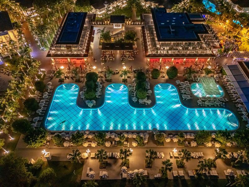 Illuminated outdoor pool in hotel complex with surrounding buildings and landscape at night.
