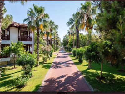 Dlážděná cesta vede zahradou s palmami a zelenými stromy vedle hotelové budovy.