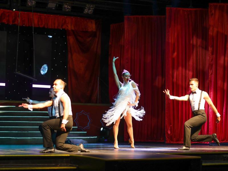 Tre ballerini sul palco con tenda rossa, due uomini in ginocchio, donna in abito bianco che danza.