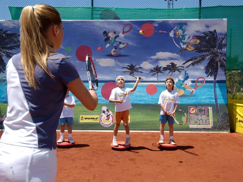 Tre bambini giocano a tennis su un campo in terra rossa mentre un allenatore li istruisce.