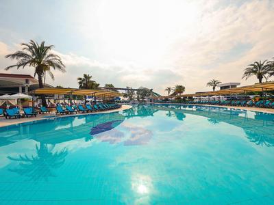 Duży basen na zewnątrz z leżakami, parasolami i palmami pod zachmurzonym niebem.