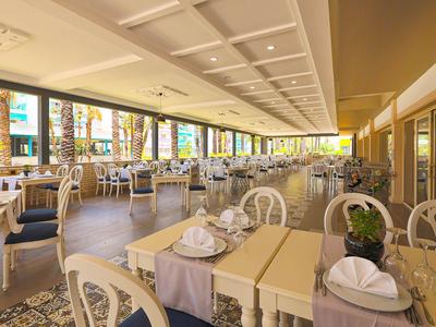 Jasna restauracja hotelowa z wieloma stołami, krzesłami i dużymi oknami z widokiem na zewnątrz.