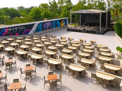 Miejsce wydarzeń na świeżym powietrzu z rzędami krzeseł, stołami i zadaszoną sceną w zielonym otoczeniu.