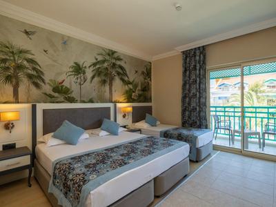 Jasny pokój hotelowy z dwoma łóżkami, stolikami nocnymi i balkonem z widokiem na palmy.