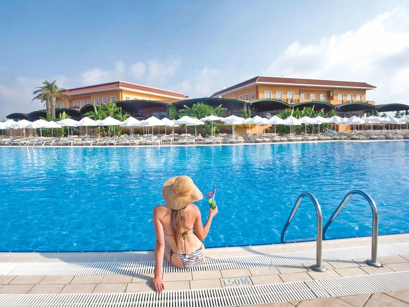 Kobieta w bikini z kapeluszem siedzi na krawędzi basenu w ośrodku, patrząc na wodę.