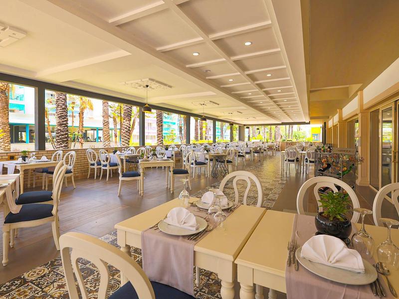 Jasna restauracja hotelowa z wieloma stołami, krzesłami i dużymi oknami z widokiem na zewnątrz.