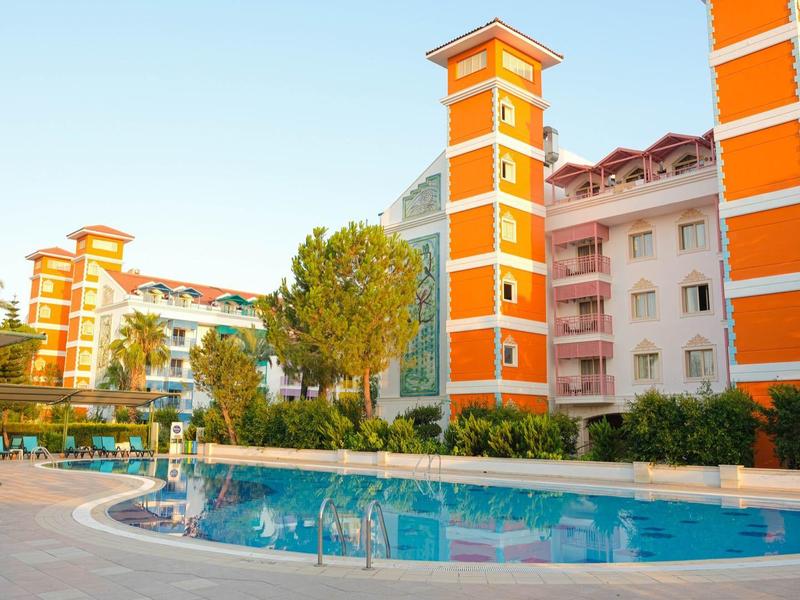 Kompleks hotelowy z pomarańczowymi budynkami i dużym odkrytym basenem w słoneczny dzień.