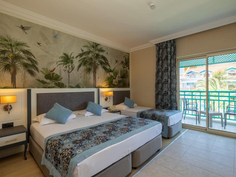 Jasny pokój hotelowy z dwoma łóżkami, stolikami nocnymi i balkonem z widokiem na palmy.